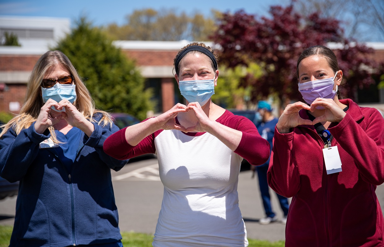 Three nurses standing outside in the sun wearing surgical face masks and holding their hands up in the shape of hearts
